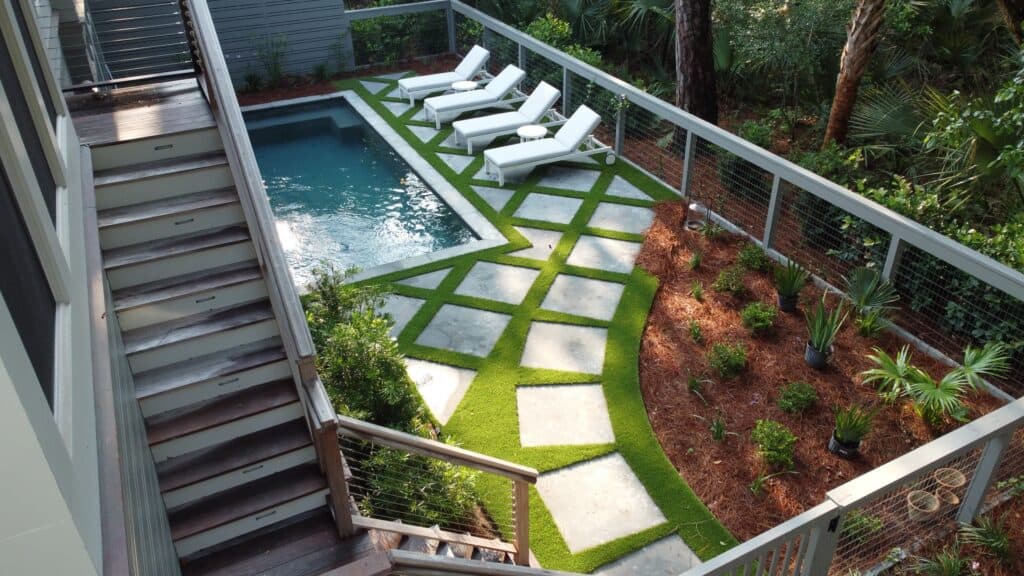 A pool with lawn chairs and a railing and artificial turf installed between pavers around the pool